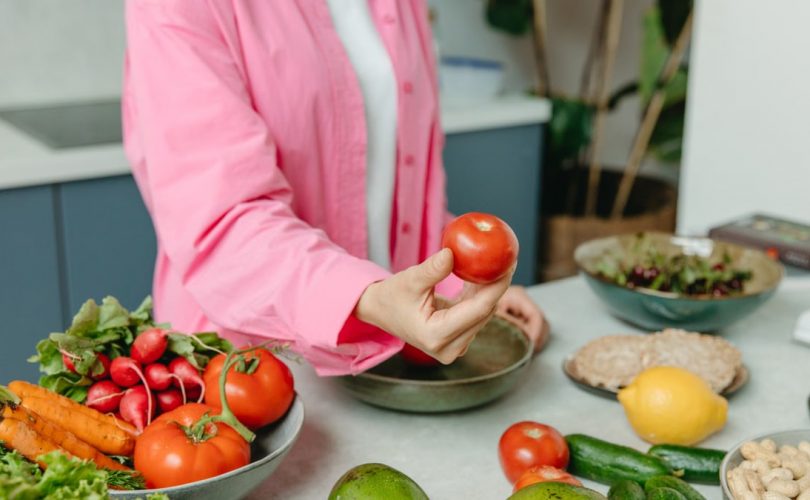 Person in pink overshirt holding a fresh tomato with various organic vegetables on a countertop.