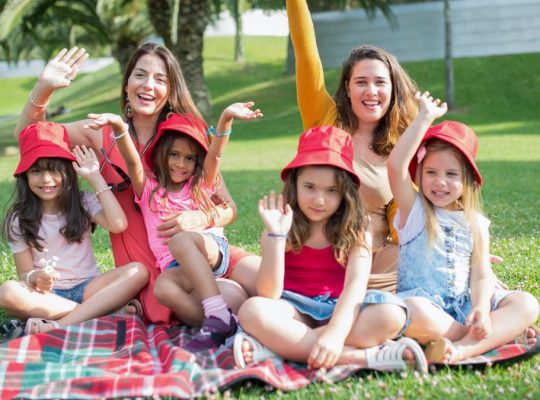 Two women and four girls enjoy a sunny picnic in Portugal, waving happily in the park.