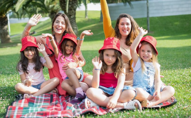 Two women and four girls enjoy a sunny picnic in Portugal, waving happily in the park.